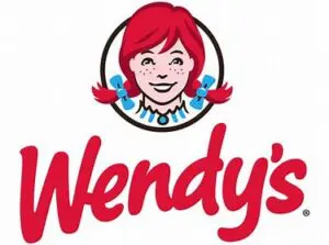 Logo of Wendy's featuring a stylized portrait of a smiling girl with red pigtails, framed in a community-based circular border, above the red, cursive text "Wendy's.