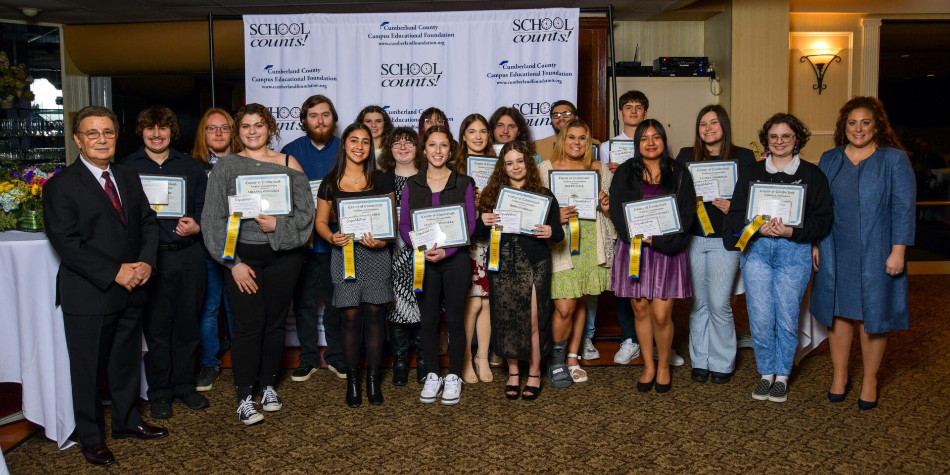 Group of students holding certificates accompanied by adults in a banquet hall with "school counts!" banners in the background.