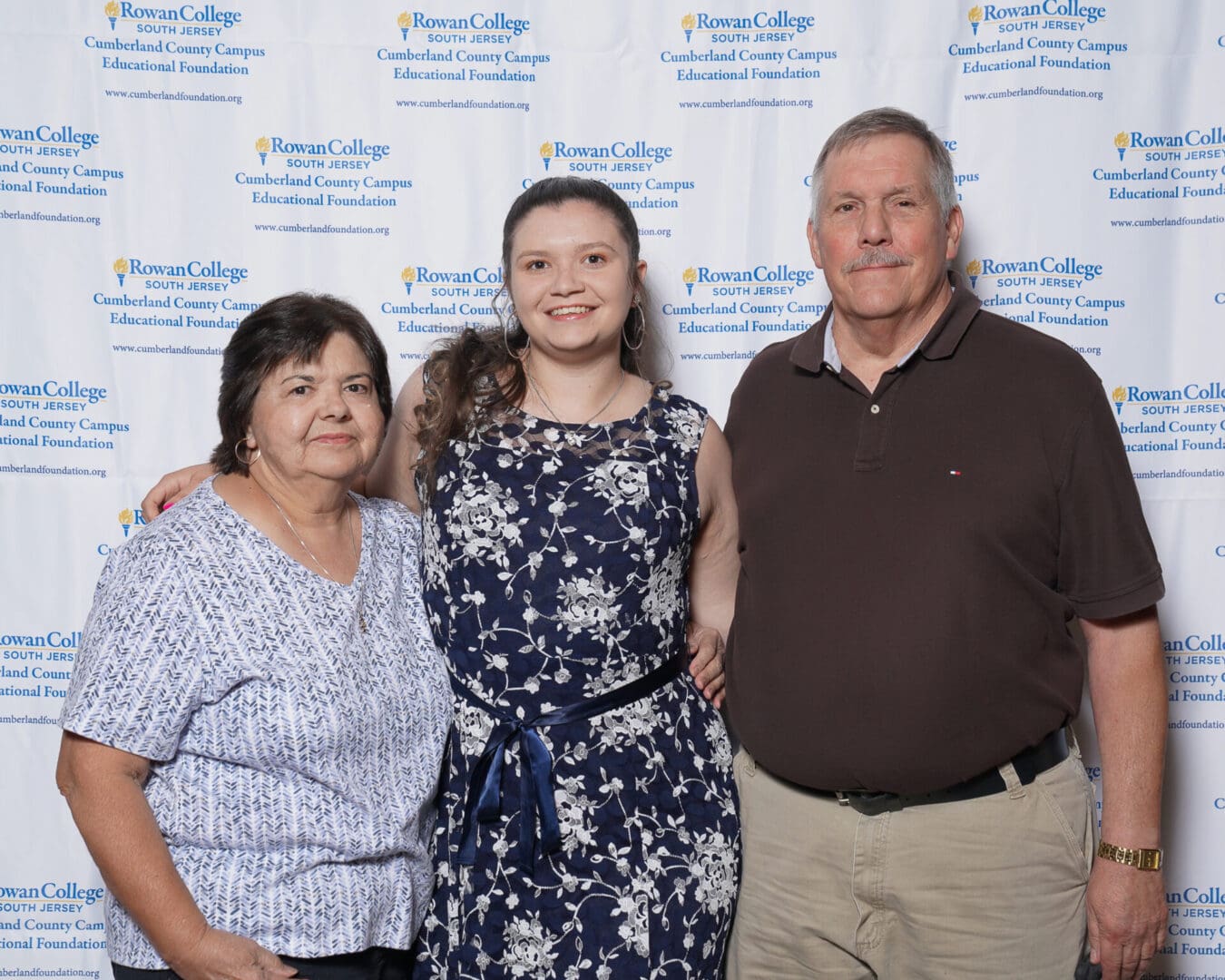 Three people posing for a photo at rowan college event, with two older adults flanking a younger woman in a floral dress, standing in front of a branded backdrop.