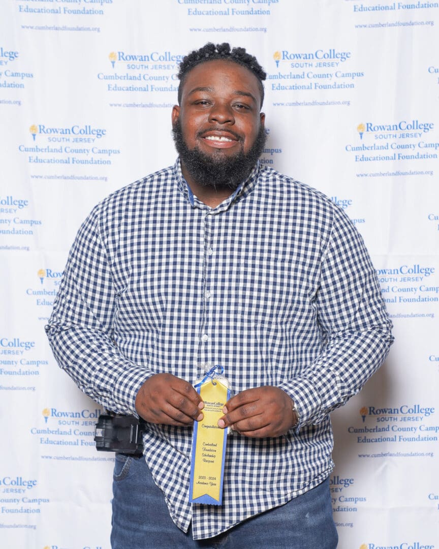 A cheerful man in a plaid shirt holding a yellow ribbon award, standing in front of a banner with educational institution logos.