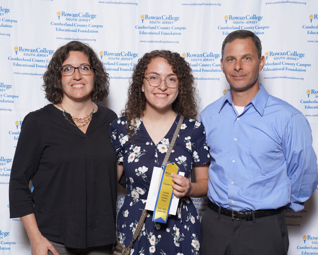 Three people posing with a certificate at a rowan college event backdrop; two women and one man, smiling, in a semi-formal setting.