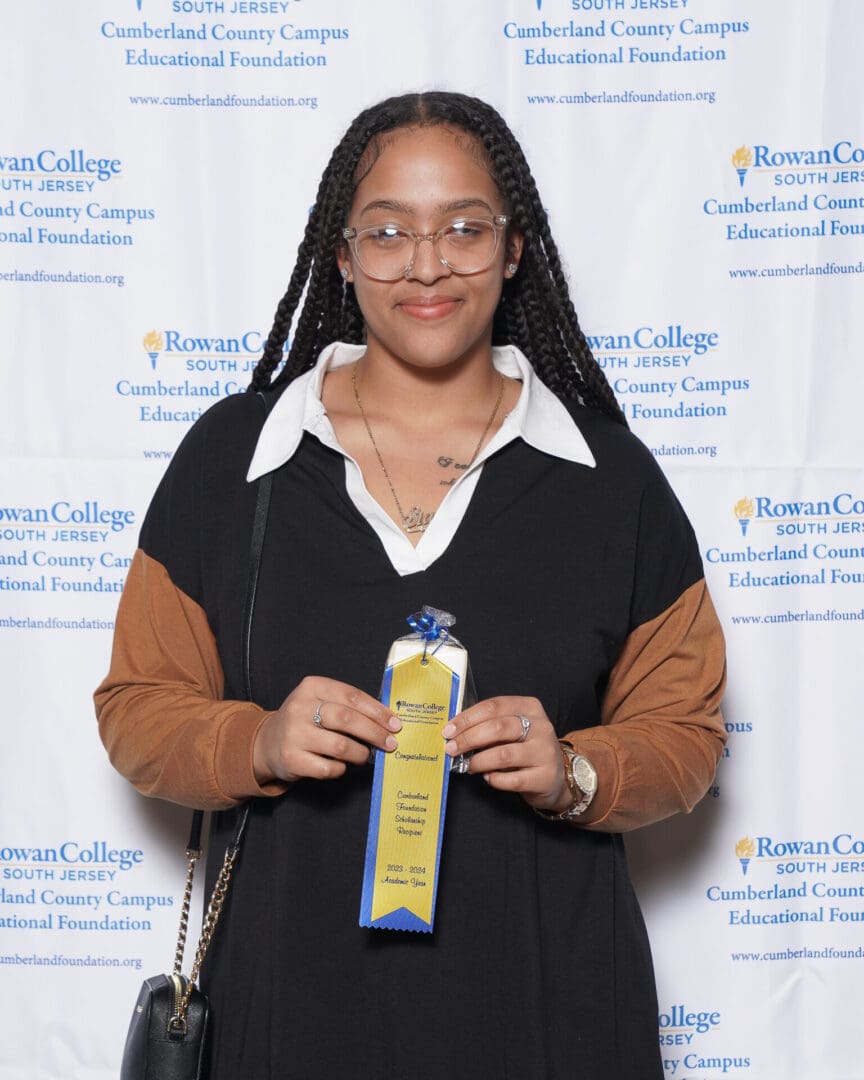 A young woman with glasses and braided hair, holding a blue award ribbon, stands in front of a cumberland foundation banner.