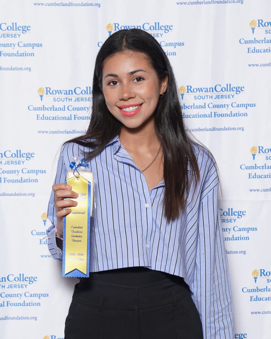 A young woman smiling at an awards ceremony, holding a gold plaque, standing in front of a banner with educational logos.