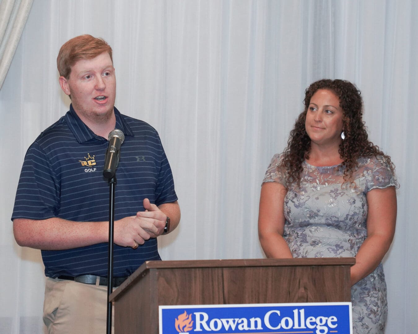 A man speaks at a podium with a rowan college logo, gesturing with his hands, as a woman stands beside him, looking on attentively.