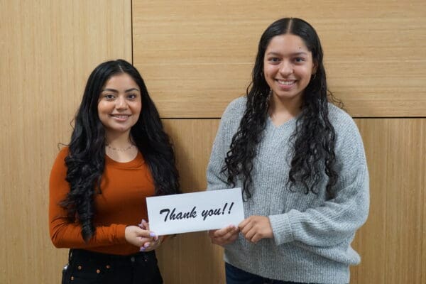 Two young women smiling at the camera, one holding a "thank you!" sign, standing in front of a wooden wall.