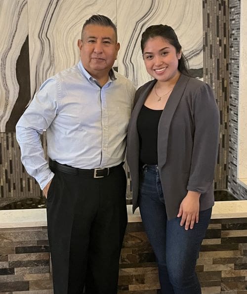 A man and a woman standing together, smiling, in front of a wavy patterned wall. both are dressed in business casual attire.