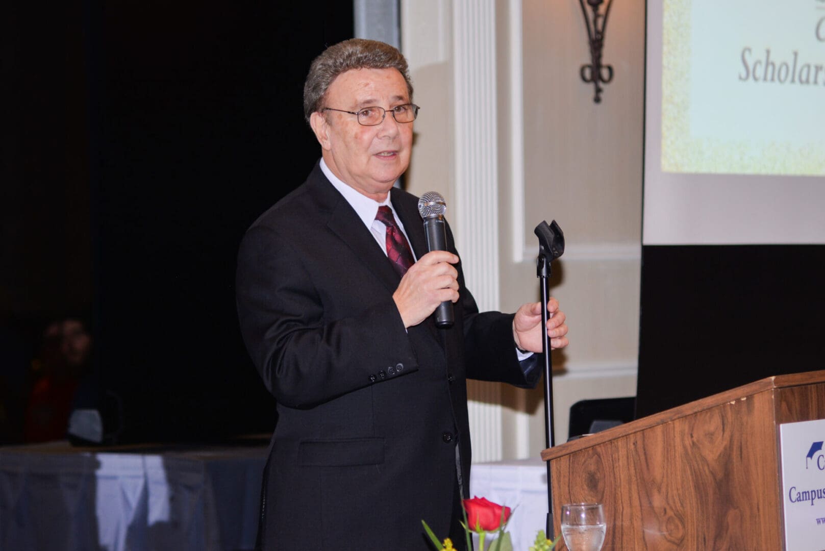 A man in a suit and tie speaking at a podium with a microphone in a dimly lit banquet hall, a banner reading "scholarship" in the background.