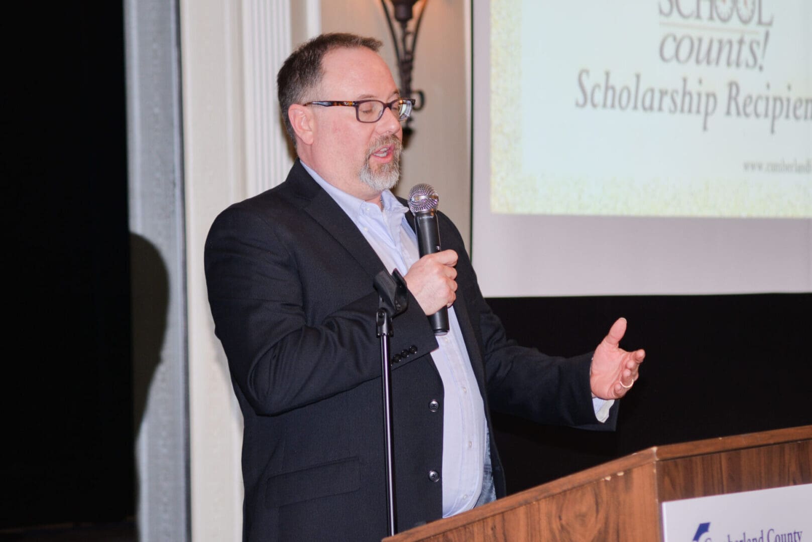 Man with glasses speaking at a podium with a microphone at a "school counts!" scholarship event, with a projected image behind him.