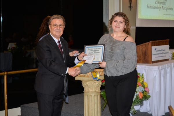 A man in a suit presenting a certificate to a woman in a gray top at a scholarship ceremony, with an audience in the background.