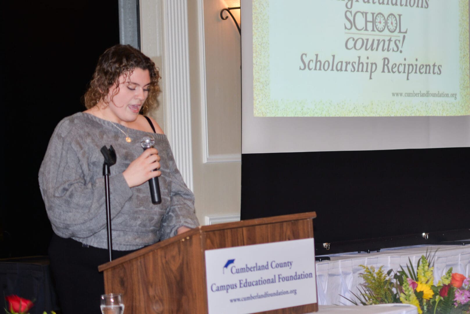 Woman speaking at a podium with a microphone, with a banner reading "cumberland county campus educational foundation" and flowers in front.