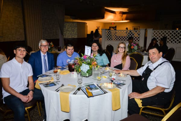 A group of six people, a mix of young adults and older men, smiling at a table with floral centerpieces in a banquet hall.