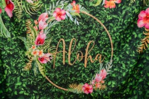 A decorative 'aloha' sign with floral accents on a lush green foliage background.