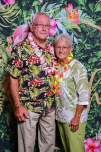 Elderly couple wearing floral outfits standing together in front of a tropical flower backdrop.