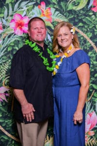 A couple, the man in a black shirt and the woman in a blue dress, adorned with floral leis, stand together smiling against a tropical floral backdrop.