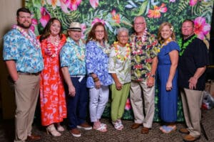 Eight adults wearing colorful tropical clothing posing together in front of a vibrant floral backdrop at an event.