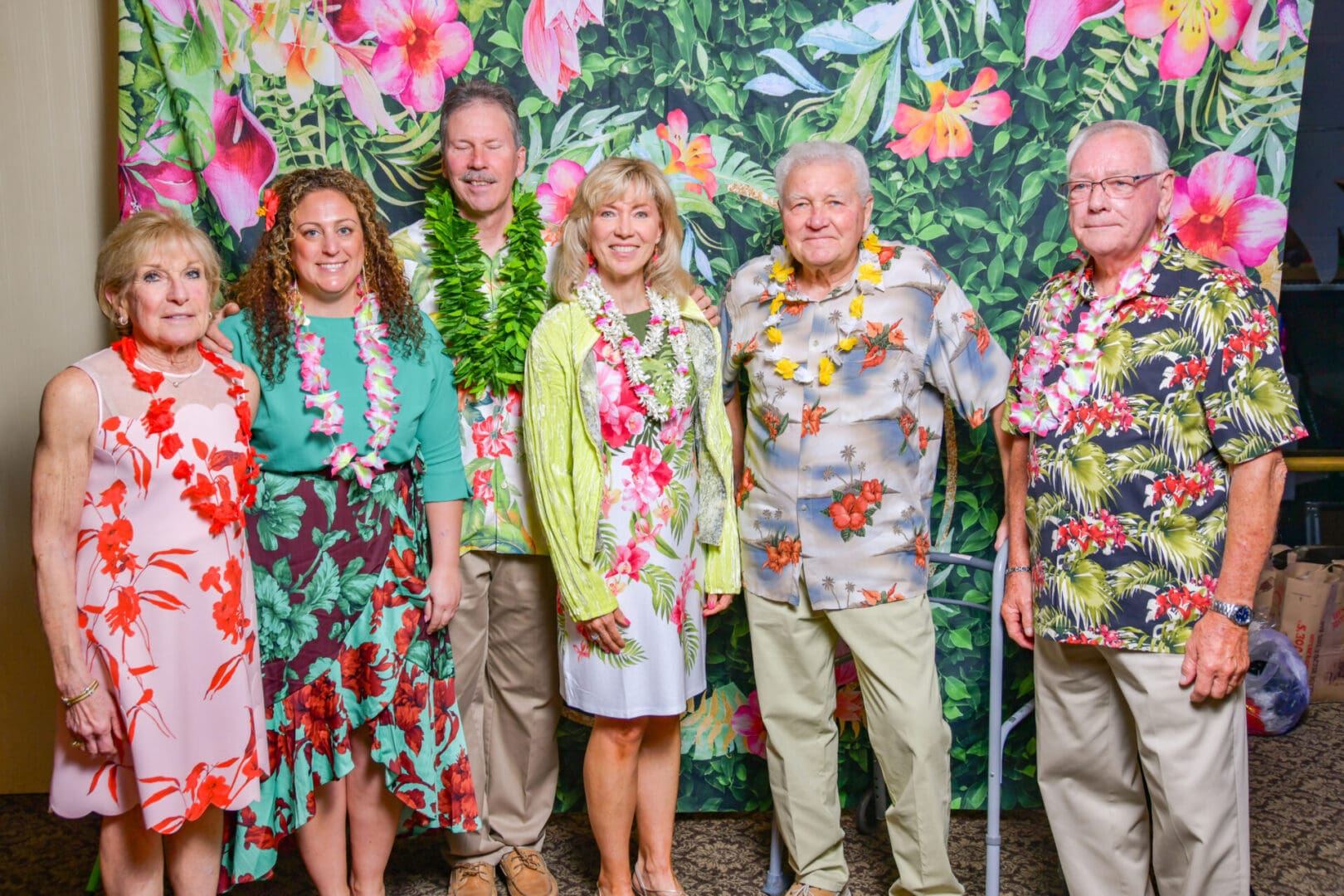 Six adults dressed in tropical-themed attire, including lei necklaces and vibrant floral shirts and dresses, posing together in front of a colorful floral backdrop.