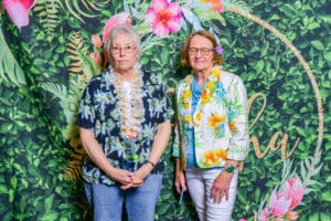 Two elderly women wearing leis stand before a floral backdrop with a golden "aloha" sign.