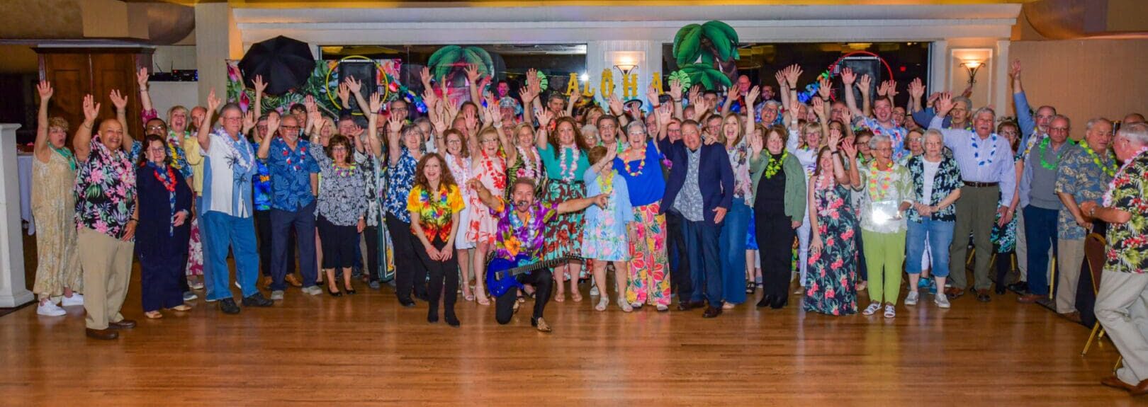 Large group of people in festive attire, some wearing hawaiian leis, joyously raising hands at a party in a decorated hall.