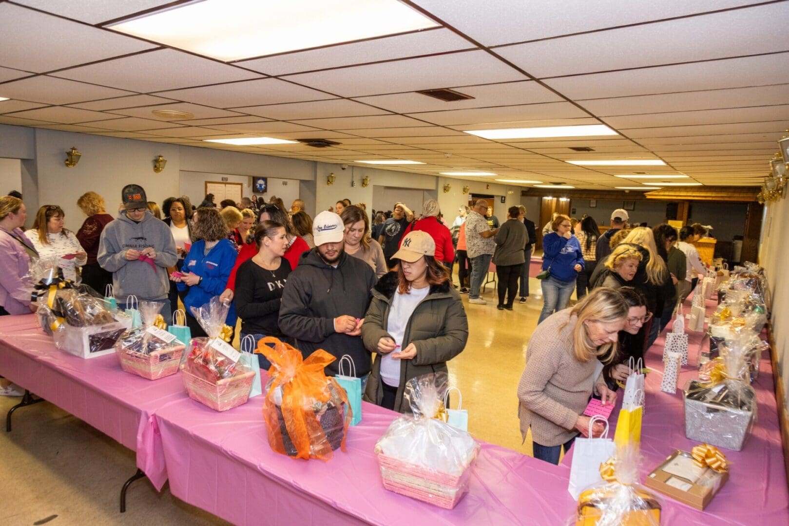 People exploring items at a charity auction event with tables covered in pink cloths and various gift baskets on display.