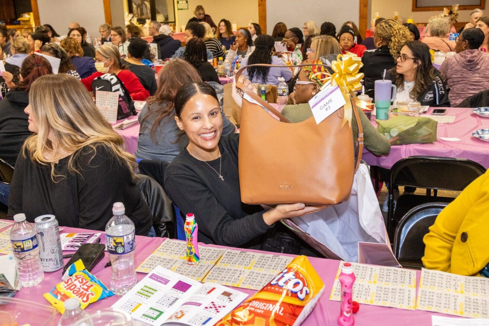 A woman smiling at a bingo event, showing off a new tan handbag with a gift bow, surrounded by other participants and snacks on the table.