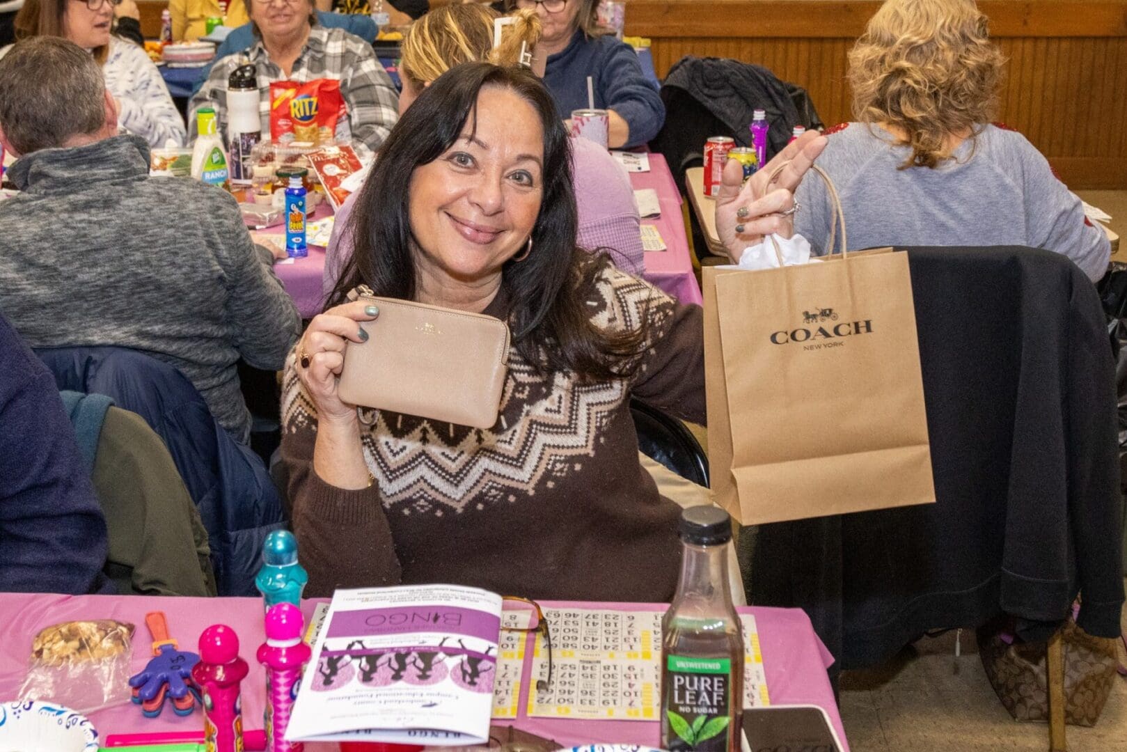 A woman smiling at a table during an event, holding up a small coach wallet with a coach shopping bag beside her.