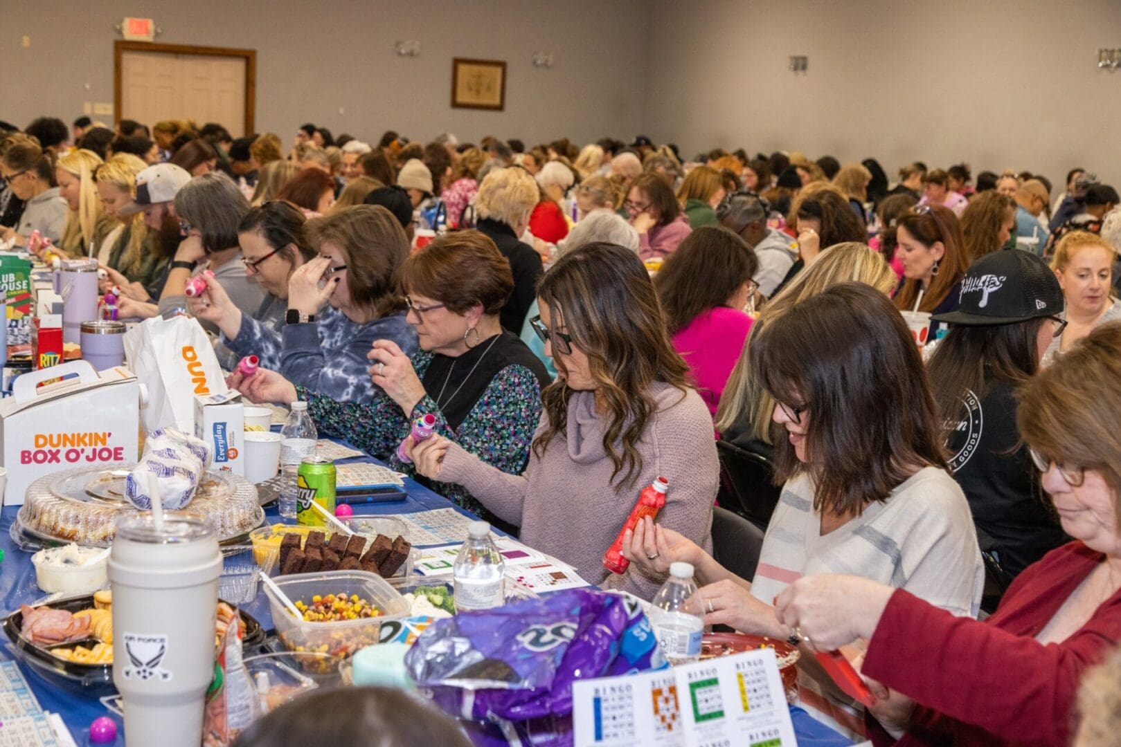 A crowded bingo hall with many people, predominantly women, focused on their bingo cards, with snacks and drinks on the tables.