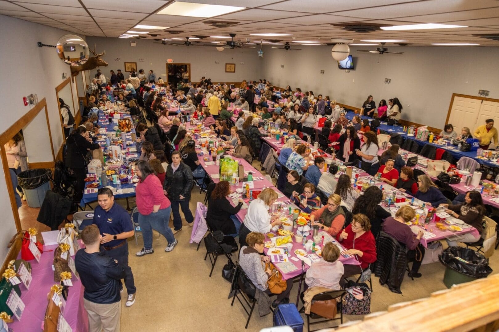 A large indoor community event with many people seated at tables, engaging in activities and conversing, surrounded by various items on the tables.