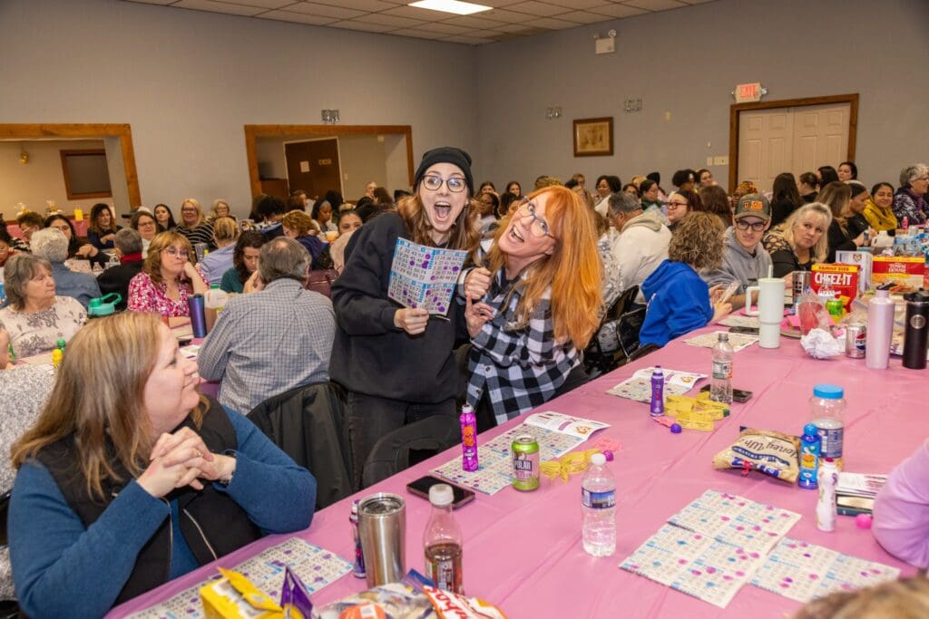 Two women excitedly celebrate a win at a crowded bingo hall, one holding up a winning bingo card.