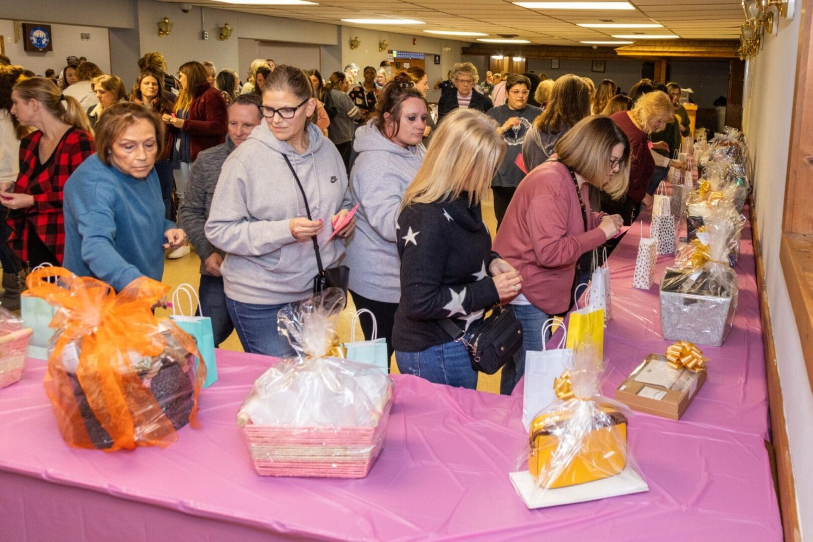 People browsing various gift baskets at a crowded indoor fundraising event.