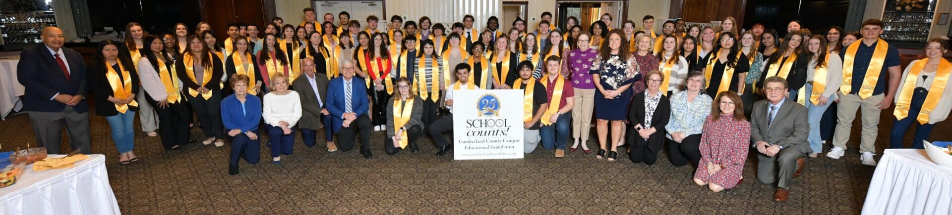 Group photo of smiling graduates with yellow stoles and a school banner.