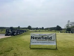A sign for Newfield Golf Course on a grassy field under a cloudy sky.