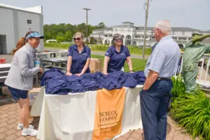 Two women in navy shirts stand behind a table with folded navy shirts, talking to an older man outdoors.