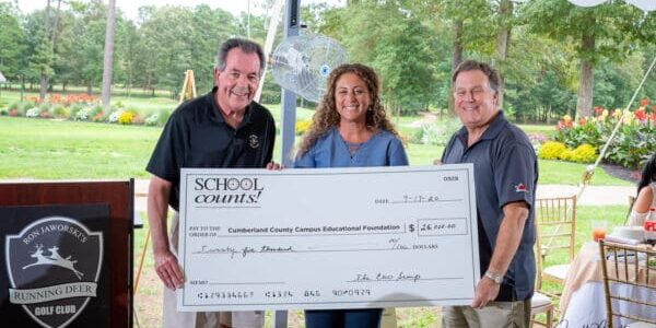 Three smiling people, two men and a woman, hold a large ceremonial check at a community-based golf event.